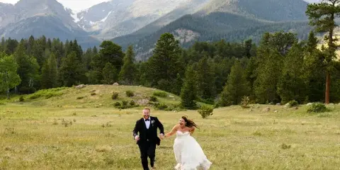 A bride and groom running hand-in-hand through a grassy meadow with a backdrop of pine trees and snow-capped mountains.