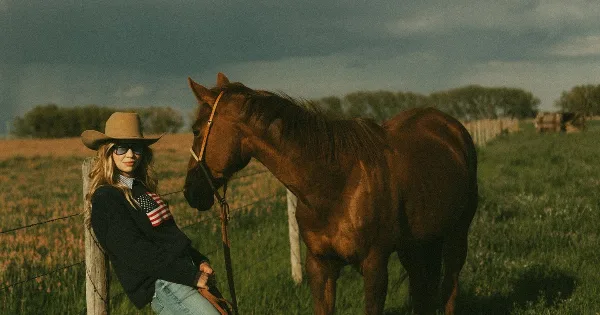 Portrait of a woman in a cowboy hat leaning against a fence post next to a brown horse in a grassy field.