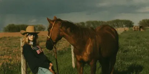 Portrait of a woman in a cowboy hat leaning against a fence post next to a brown horse in a grassy field.
