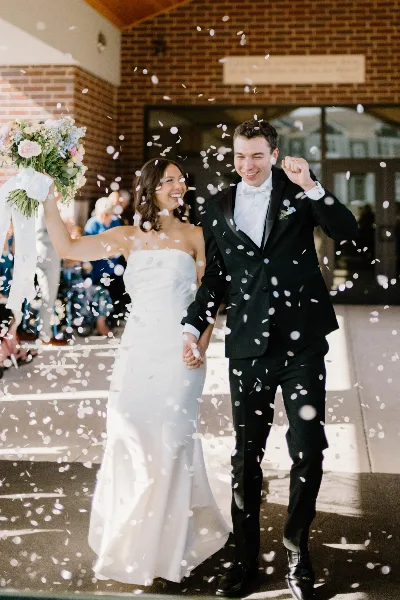 A bride and groom walking hand-in-hand while flower petals fall around them.