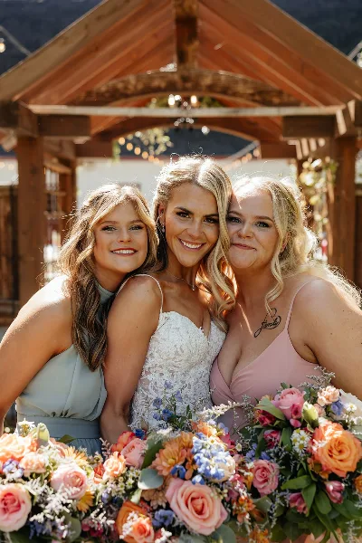 A bride and two bridesmaids posing together with styled hair and makeup in front of a wooden wedding arch.