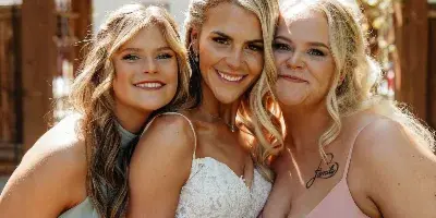 A bride and two bridesmaids posing together with styled hair and makeup in front of a wooden wedding arch.