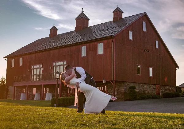 A bride and groom kissing in front of a large red barn wedding venue.