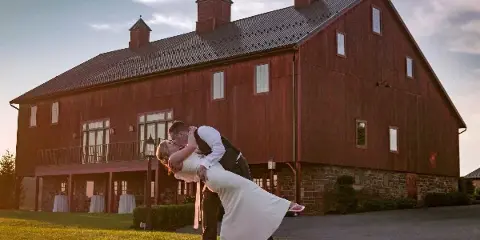 A bride and groom kissing in front of a large red barn wedding venue.