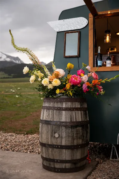 A vibrant floral arrangement in a wooden barrel at an outdoor event with mountains in the background.