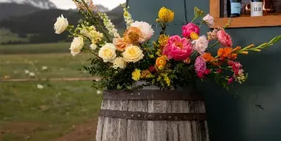 A vibrant floral arrangement in a wooden barrel at an outdoor event with mountains in the background.
