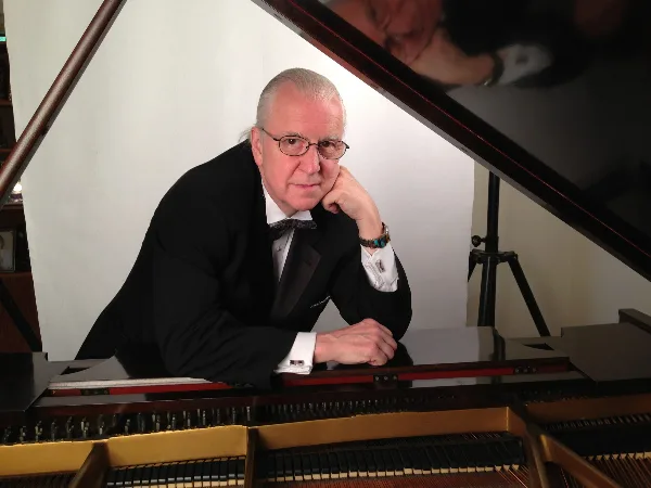 A man in a black tuxedo and bow tie leaning against a grand piano with the lid open.