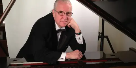 A man in a black tuxedo and bow tie leaning against a grand piano with the lid open.