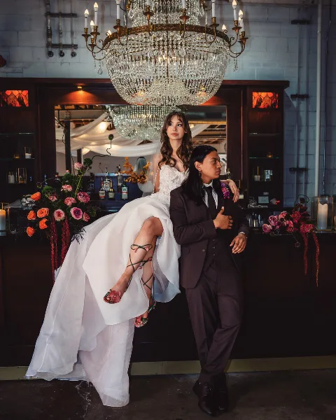 A bride in a white gown and a groom in a dark suit posing together at a bar with a large crystal chandelier and floral arrangements.