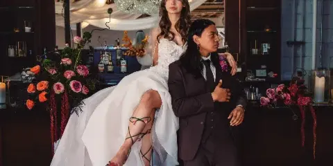 A bride in a white gown and a groom in a dark suit posing together at a bar with a large crystal chandelier and floral arrangements.