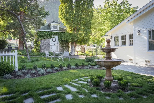 A landscaped garden at a white building featuring a stone fountain, manicured lawn, and a small ivy-covered structure.