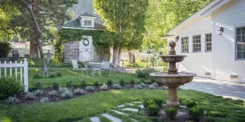 A landscaped garden at a white building featuring a stone fountain, manicured lawn, and a small ivy-covered structure.