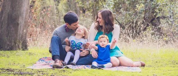 A family of four sitting on a patterned blanket on a grassy lawn, with the parents interacting affectionately with their two young children.