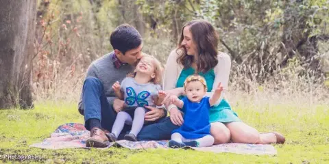 A family of four sitting on a patterned blanket on a grassy lawn, with the parents interacting affectionately with their two young children.