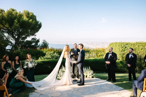 A bride and groom exchanging vows during an outdoor wedding ceremony with an officiant and wedding party present.