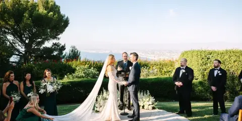 A bride and groom exchanging vows during an outdoor wedding ceremony with an officiant and wedding party present.