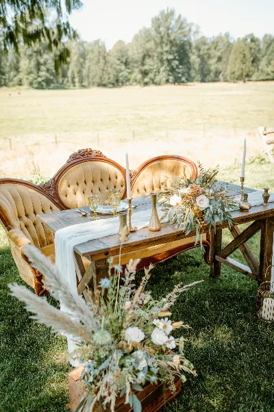 An outdoor wedding table setting with antique chairs, a wooden table, and floral centerpieces.