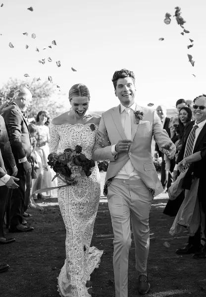 A black and white photograph of a bride and groom walking down the aisle during a wedding ceremony, surrounded by guests and falling petals.