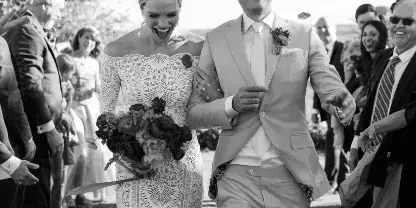 A black and white photograph of a bride and groom walking down the aisle during a wedding ceremony, surrounded by guests and falling petals.