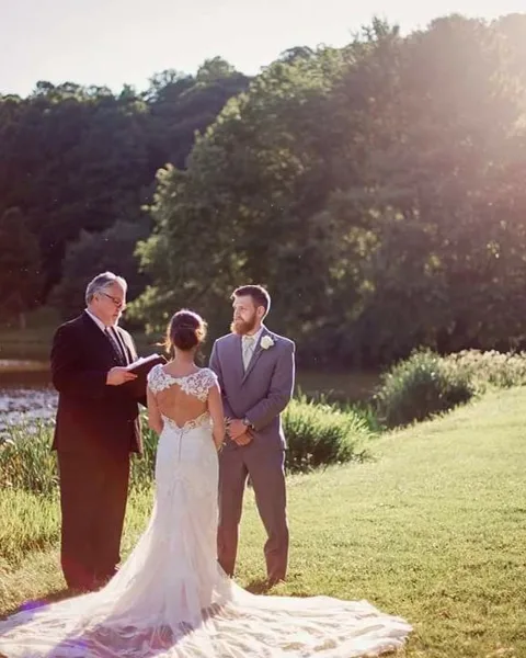 A wedding officiant in a dark suit officiating an outdoor ceremony for a bride and groom.