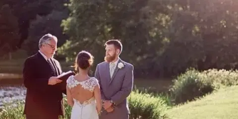 A wedding officiant in a dark suit officiating an outdoor ceremony for a bride and groom.