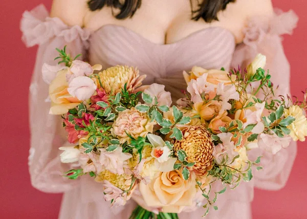 A bride in a pink gown holding a large, lush bouquet of peach roses, yellow dahlias, and greenery against a pink background.