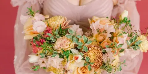A bride in a pink gown holding a large, lush bouquet of peach roses, yellow dahlias, and greenery against a pink background.