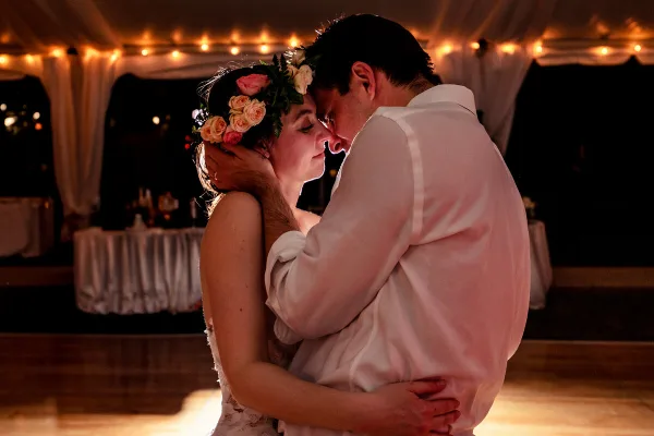 A bride wearing a floral crown and a groom in a white shirt share an intimate embrace during a wedding reception.