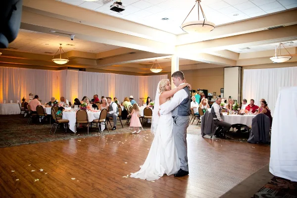A bride and groom dancing in a large banquet hall with guests seated at tables in the background.