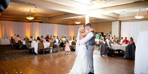 A bride and groom dancing in a large banquet hall with guests seated at tables in the background.