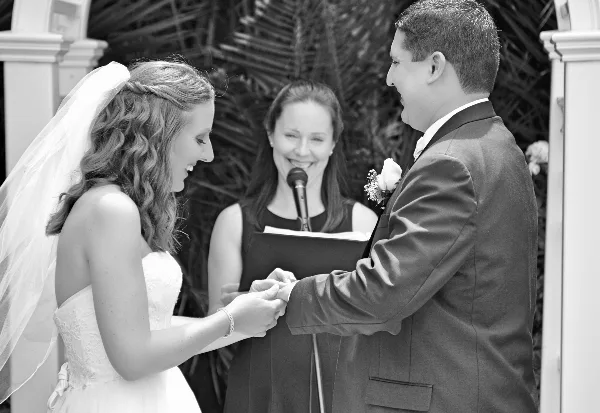 A black and white photograph of a bride and groom exchanging rings during a wedding ceremony, with an officiant standing between them holding a microphone and a book.