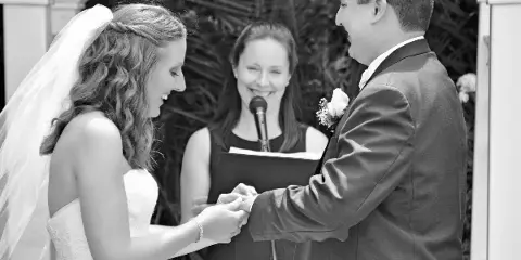 A black and white photograph of a bride and groom exchanging rings during a wedding ceremony, with an officiant standing between them holding a microphone and a book.