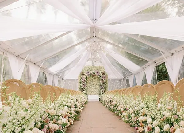 A wedding ceremony setup inside a clear tent with white draping, chandeliers, and floral arrangements.