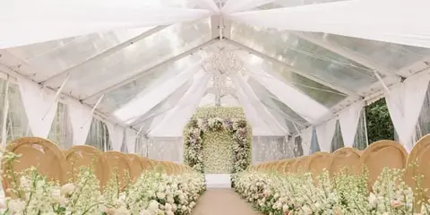 A wedding ceremony setup inside a clear tent with white draping, chandeliers, and floral arrangements.