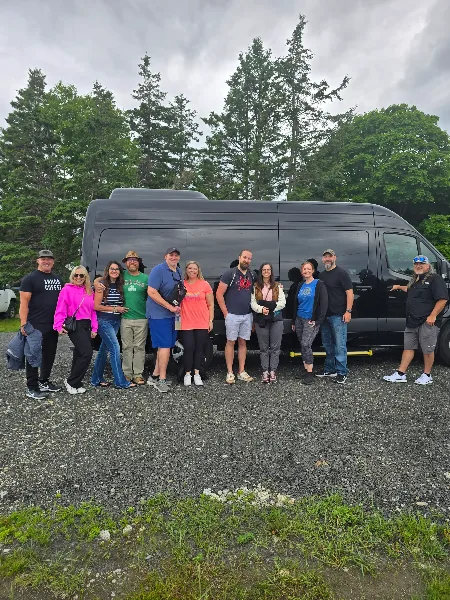 A group of people standing in front of a black Sprinter van in a gravel parking area.