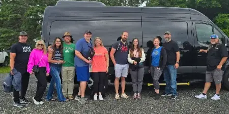 A group of people standing in front of a black Sprinter van in a gravel parking area.