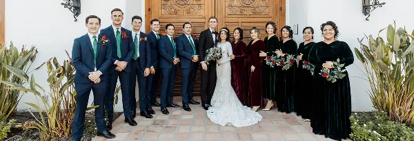 A professional wedding photograph of a bride and groom standing with their wedding party in front of a wooden door.