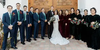 A professional wedding photograph of a bride and groom standing with their wedding party in front of a wooden door.