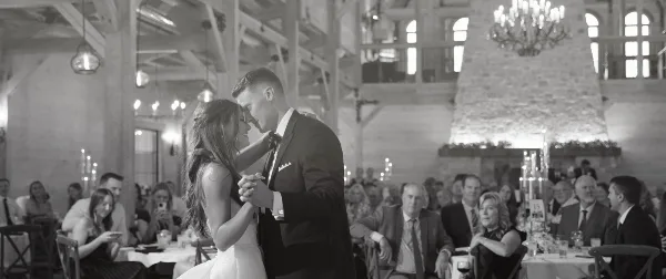 A black and white photograph of a bride and groom dancing at a wedding reception in a rustic venue with guests seated at tables in the background.
