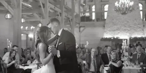 A black and white photograph of a bride and groom dancing at a wedding reception in a rustic venue with guests seated at tables in the background.