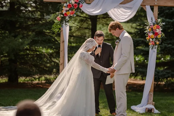A bride and groom exchanging vows under a floral and white-draped wooden arch in an outdoor setting.