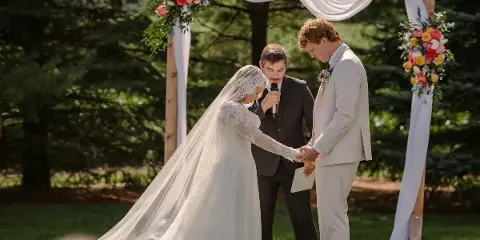 A bride and groom exchanging vows under a floral and white-draped wooden arch in an outdoor setting.