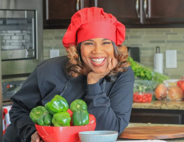 A smiling woman wearing a red chef's hat and black apron poses in a kitchen holding a red bowl filled with green bell peppers.