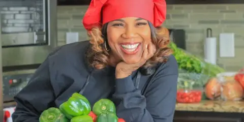 A smiling woman wearing a red chef's hat and black apron poses in a kitchen holding a red bowl filled with green bell peppers.
