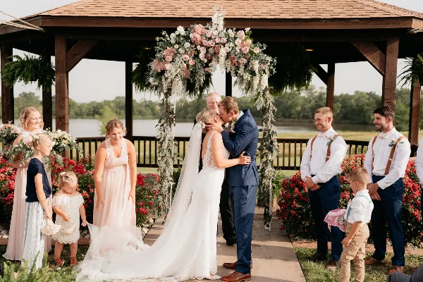 A bride and groom kissing during a wedding ceremony in a wooden gazebo with a lake view.
