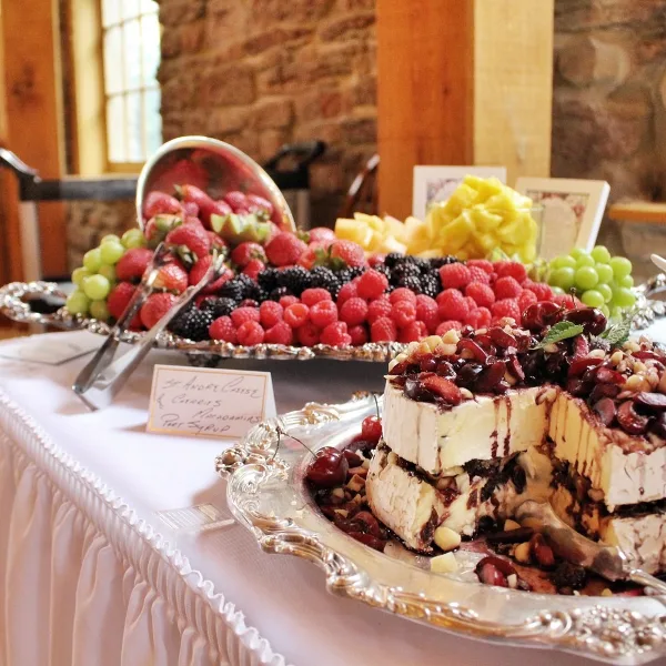 A catering table featuring a silver platter of assorted fresh berries and a layered cheesecake dessert topped with cherries and nuts.