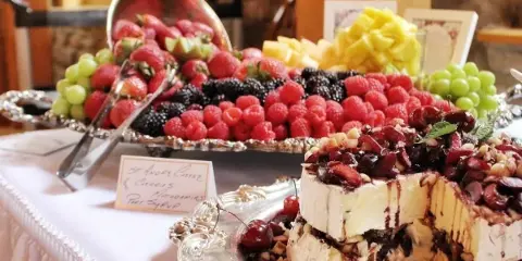 A catering table featuring a silver platter of assorted fresh berries and a layered cheesecake dessert topped with cherries and nuts.