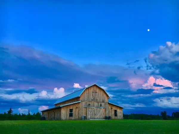 A wooden gambrel barn with a metal roof sitting on a green lawn under a blue sky with clouds and a moon.