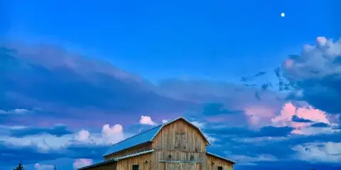 A wooden gambrel barn with a metal roof sitting on a green lawn under a blue sky with clouds and a moon.