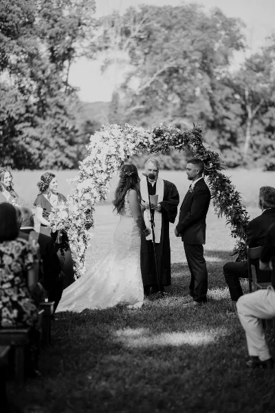 Black and white photograph of an outdoor wedding ceremony featuring a bride and groom standing under a floral arch with an officiant and seated guests.